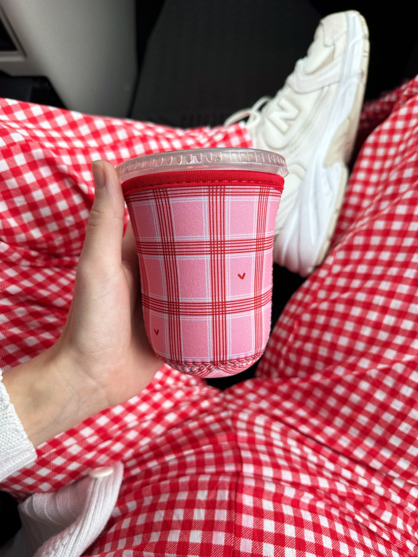 Holding an iced coffee with a pink and red checkered sleeve with hearts for valentines day against gingham pants