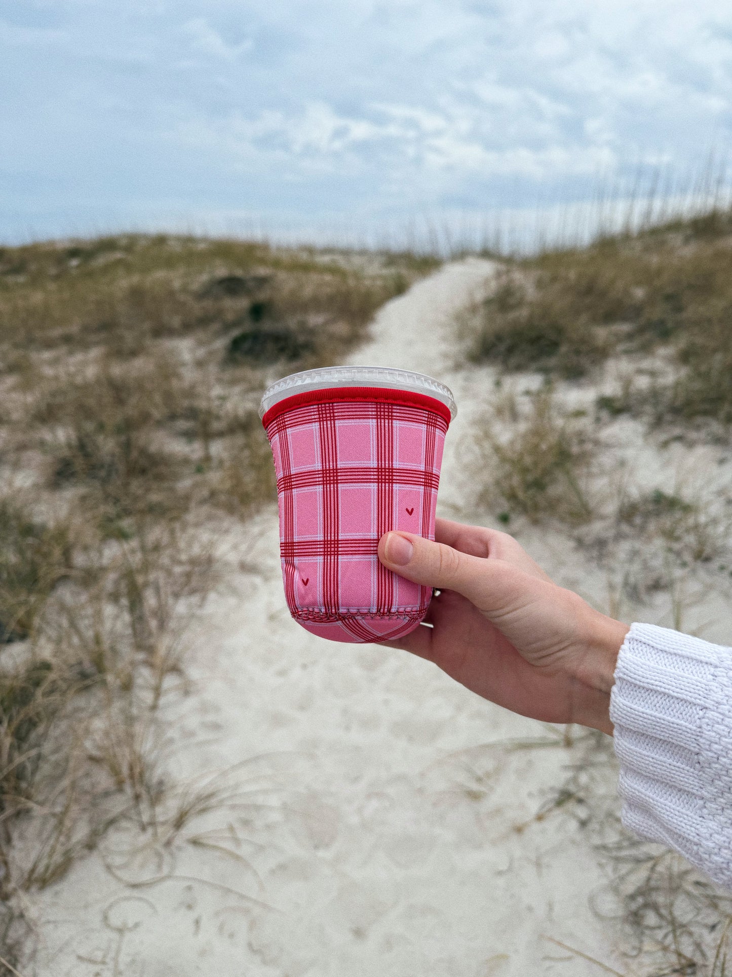Hand holding pink and red checkered iced coffee sleeve with hearts for valentines day at the beach