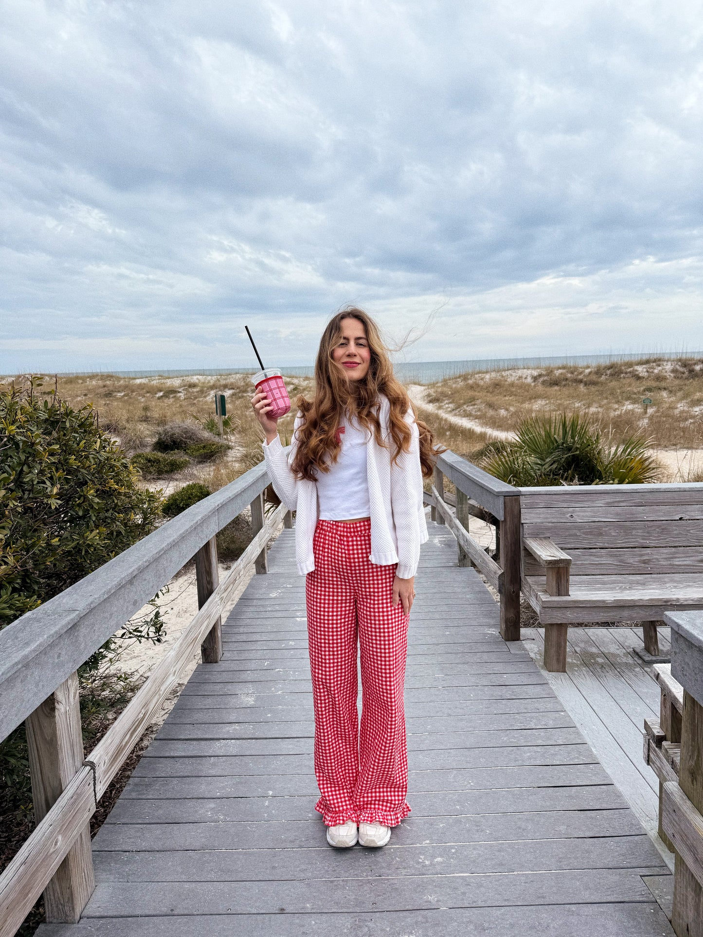 Girl holding pink and red checkered iced coffee sleeve with hearts for valentines day at the beach with a bow shirt and gingham pants onion a pinterest style photo