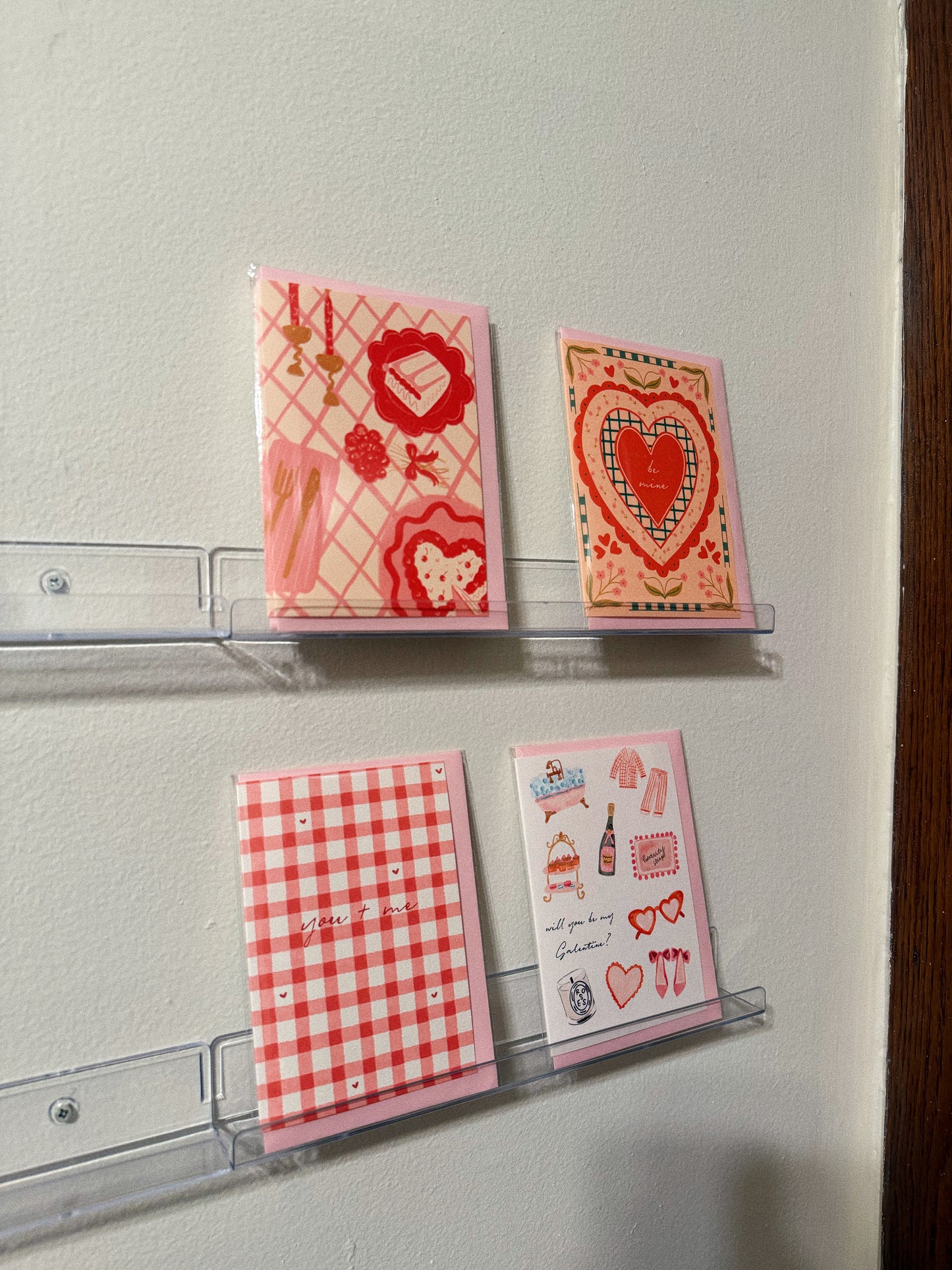 4 greeting cards for valentines day on clear shelves being displayed, one with a table setting one with a red heart that says be mine, one that says you + me and one that says happy galentines day with girly watercolor doodles.
