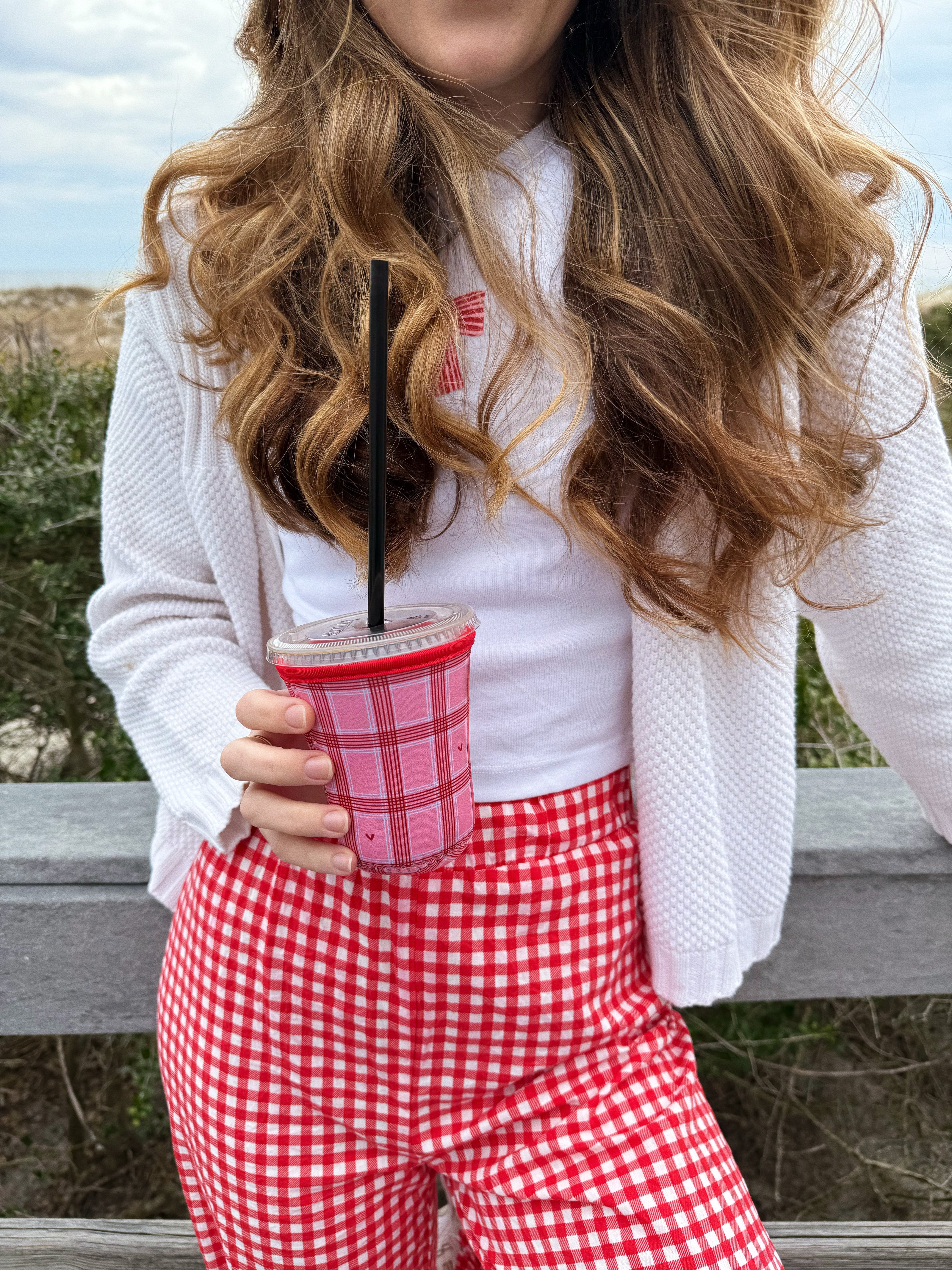 Girl holding pink and red checkered iced coffee sleeve with hearts for valentines day at the beach with a bow shirt and gingham pants on